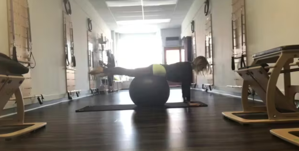 Woman doing Pilates exercises on a yoga mat with a Pilates ball. She is in a Pilates studio with Pilates equipment.