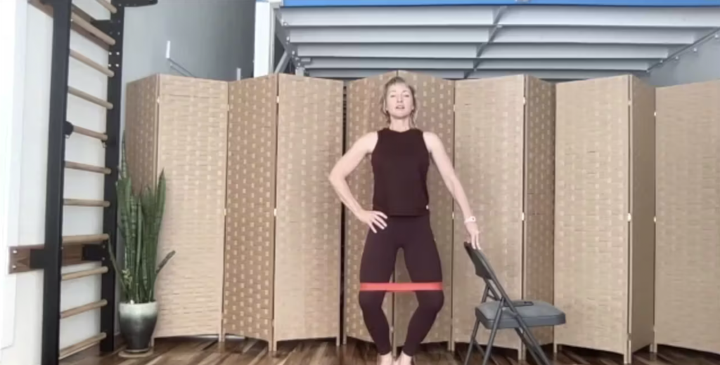 Woman doing Pilates exercises standing with a chair. She has a loop band around her thighs.