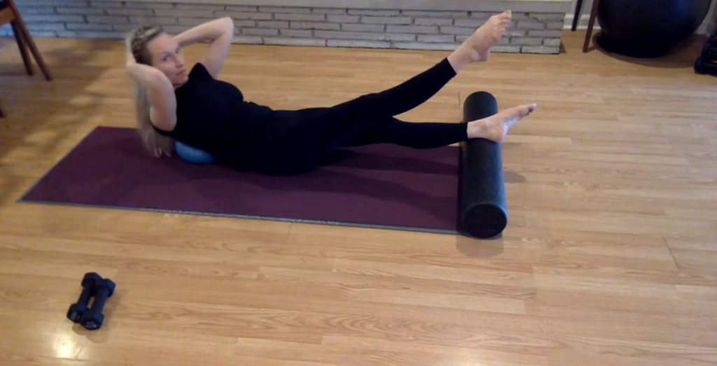 Woman doing Pilates exercises on the Pilates mat. There is a foam roller, Pilates ball, and weights around her.