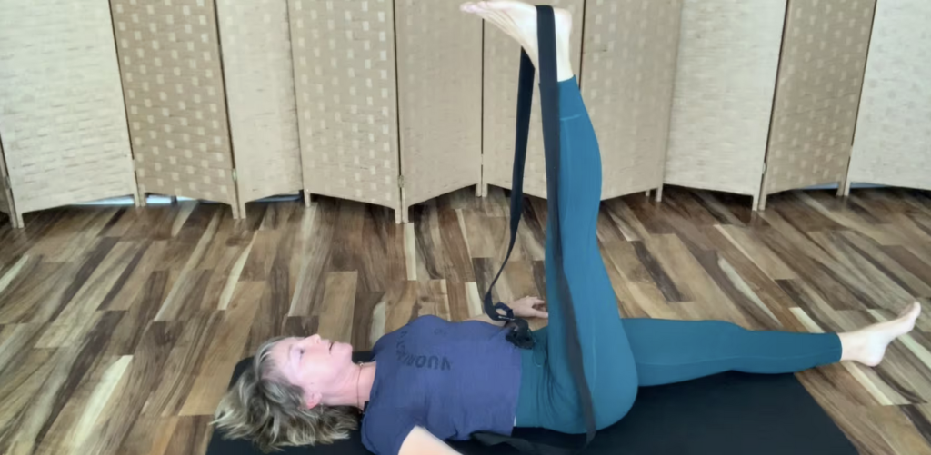 Woman doing Pilates mat exercises on the floor lying on her back with one leg up and yoga strap