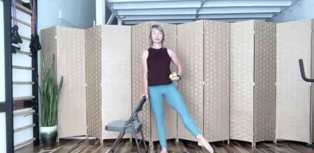 Woman in virtual Pilates and barre class holding weights and standing next to a chair.