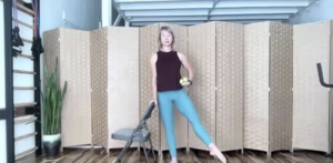 Woman in virtual Pilates and barre class holding weights and standing next to a chair.