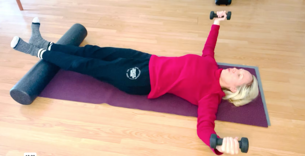 Woman lying on a Pilates mat doing arm exercises with weights and leg exercises with a foam roller.