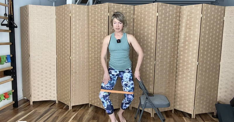 Woman standing next to a chair with a loop band around her legs doing PIlates exercises.