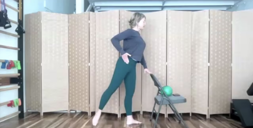 Woman standing with a chair extending one leg back for barre and Pilates exercises.