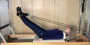 Woman doing Pilates on Pilates reformer. Her feet are in the Pilates straps and she is lying on the reformer.