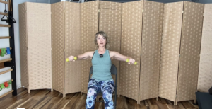 Woman seated in a chair holding weights doing Pilates arm exercises.