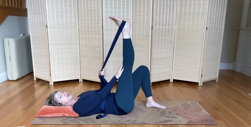 Woman lying on Pilates mat with one leg extended and yoga strap.