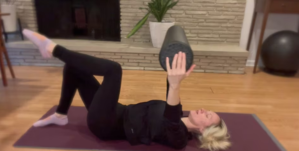 Woman doing Pilates exercise on a yoga mat holding a foam roller with one leg up.