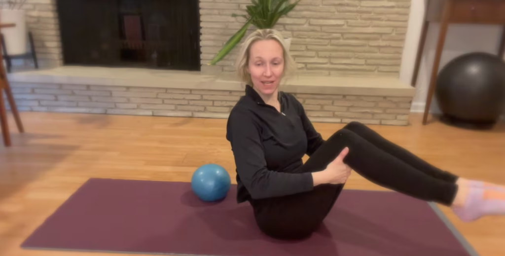 Woman seated on a Pilates mat doing a Pilates teaser exercise.