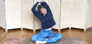 Woman seated on a Pilates mat stretching her tricep muscle.