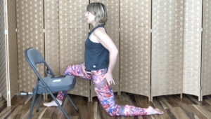 Woman kneeling on one knee doing Pilates exercises next to a chair.