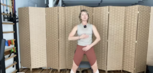 Woman standing doing barre exercises on a Pilates mat in front of a chair.