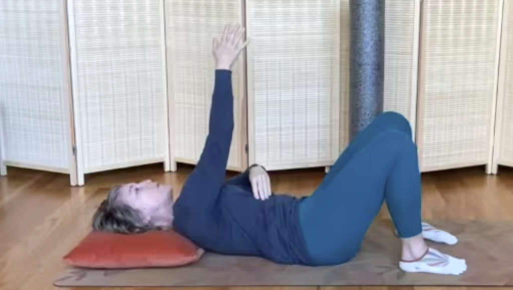 Woman lying on the floor doing Pilates exercises with one arm extended up.